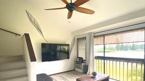 A modern living room with a wooden floor, a ceiling fan, a flat-screen TV, a sofa, a coffee table, and a balcony with a view of greenery.