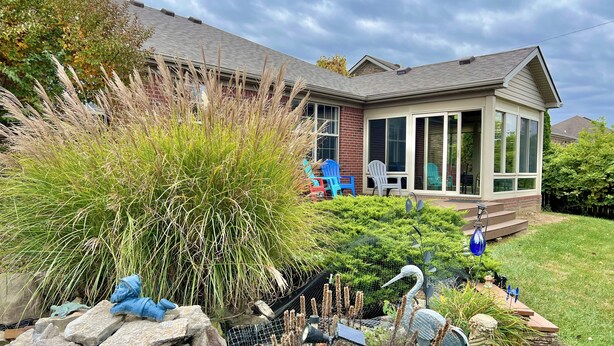 Ein Haus mit überdachter Veranda, einem Garten mit verschiedenen Pflanzen und Skulpturen sowie einer Ziegelaußenwand.