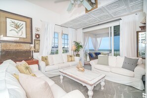 A spacious living room with white furniture, a large window with a view, and a ceiling with intricate patterns.