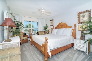 A bedroom with a wooden bed, wicker furniture, a ceiling fan, and a view of palm trees through the window.