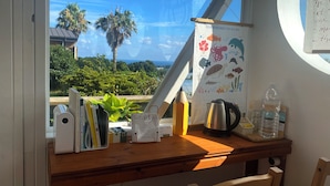 Escritorio con libros, una tetera y una botella de agua, con vista a un paisaje con palmeras y cielo despejado.