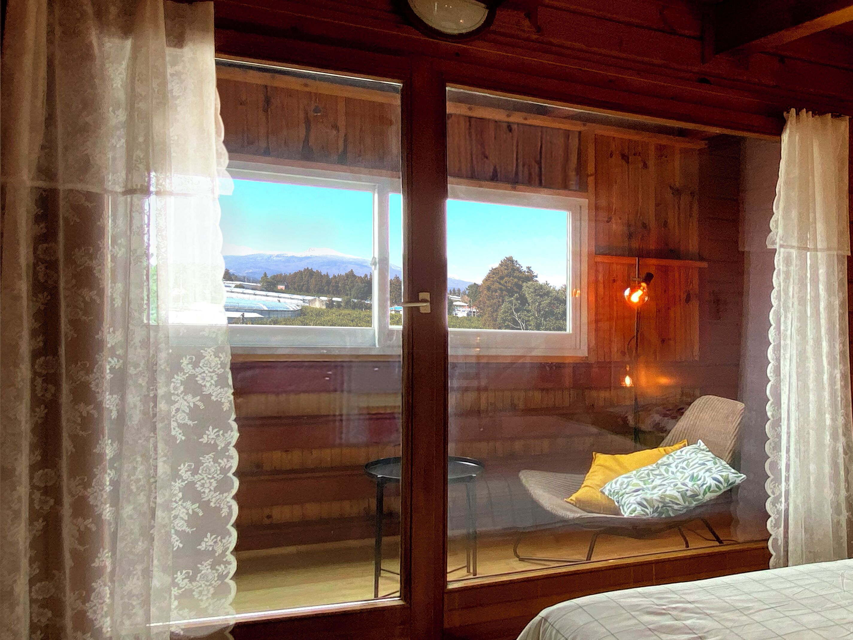 A wooden cabin room with a bed, a chair, a table, and a view of a mountain and trees through a window.