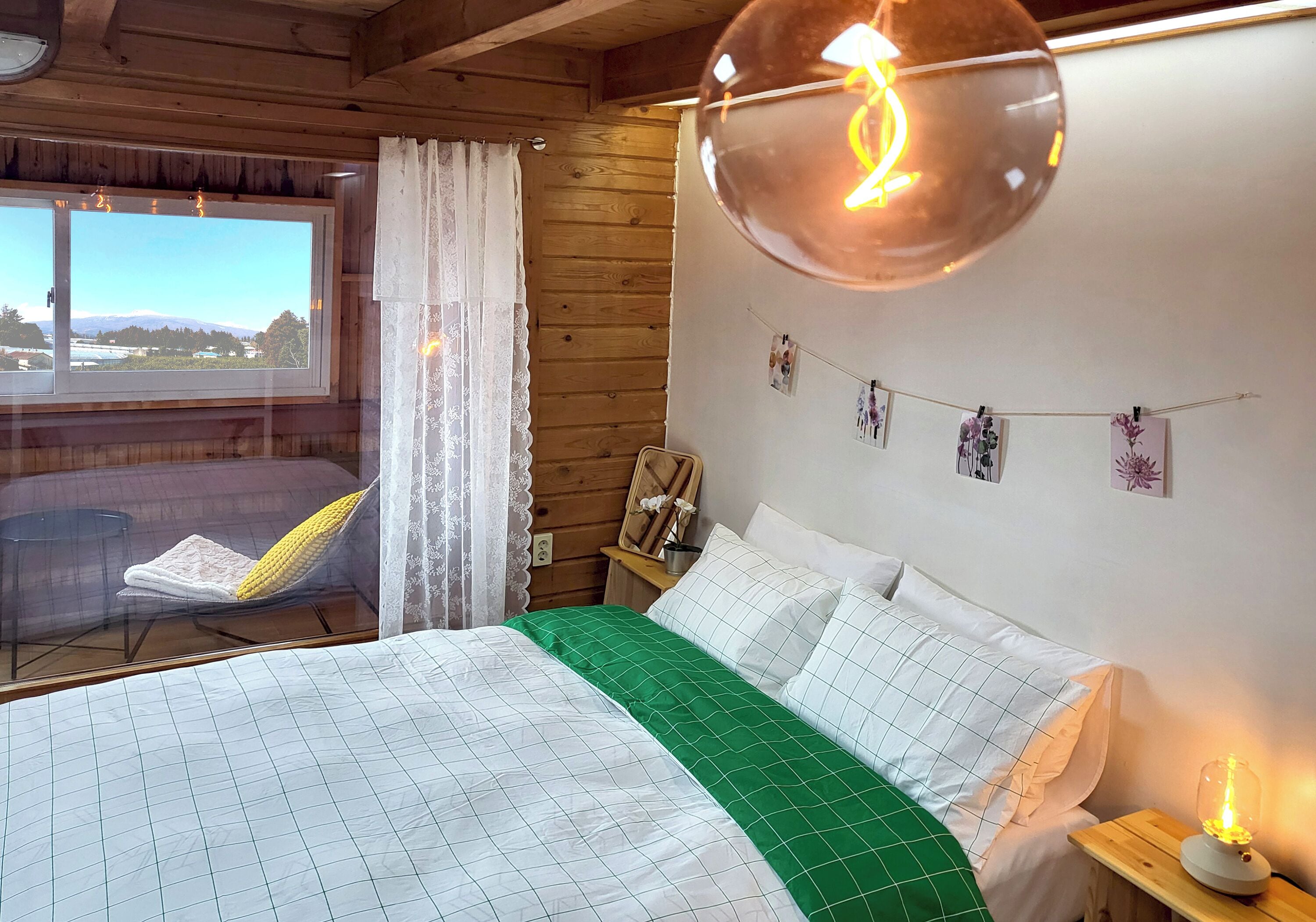 A bedroom with a bed, a bedside table with a lamp, a window with curtains, and a view of the outdoors.