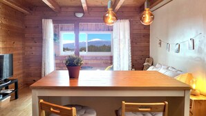 Interior de una cabaña de madera con una mesa de comedor, dos sillas, una planta en maceta, un televisor y vistas a las montañas a través de las ventanas.