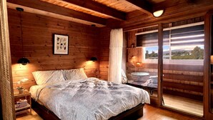 Un dormitorio de cabaña de madera con cama, lámparas de noche, una impresión enmarcada de motivos botánicos y un ventanal con vistas.