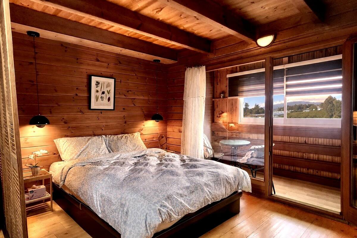 A wooden cabin bedroom with a bed, bedside lamps, a framed botanical print, and a large window with a view.