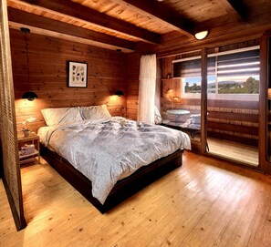 A wooden cabin bedroom with a bed, bedside lamps, a framed botanical print, and a large window with a view.
