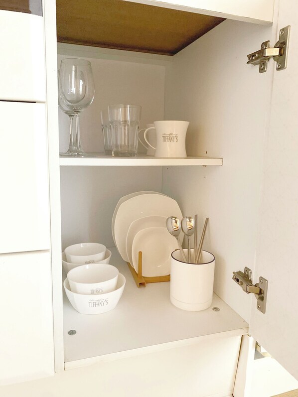 A white cabinet with shelves containing white bowls, a white mug, and a set of silver spoons.