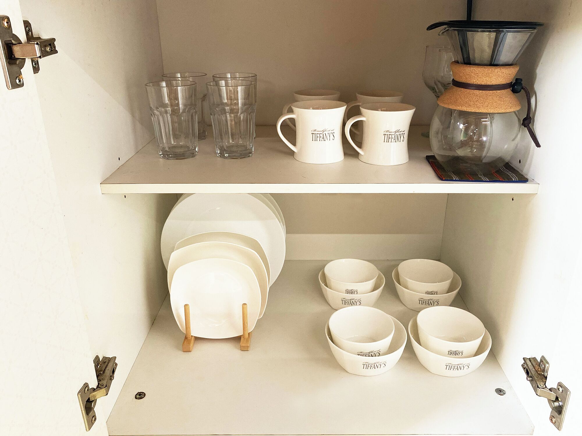 Estante con vasos transparentes, tazas y boles blancos con la marca 'TIFFANY'S'.
