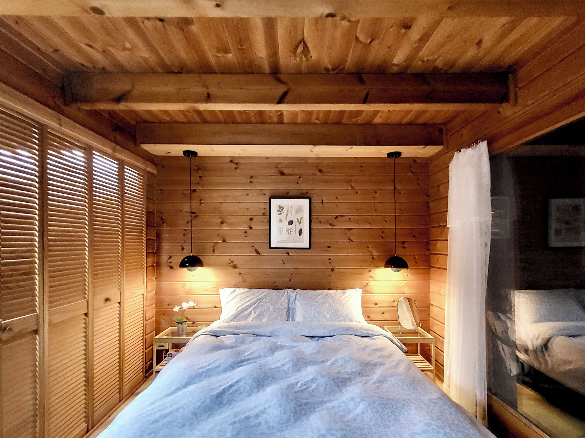 A wooden cabin room with a bed, two bedside tables, a framed picture on the wall, and a window with blinds.