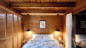 A wooden cabin room with a bed, two bedside tables, a framed picture on the wall, and a window with blinds.