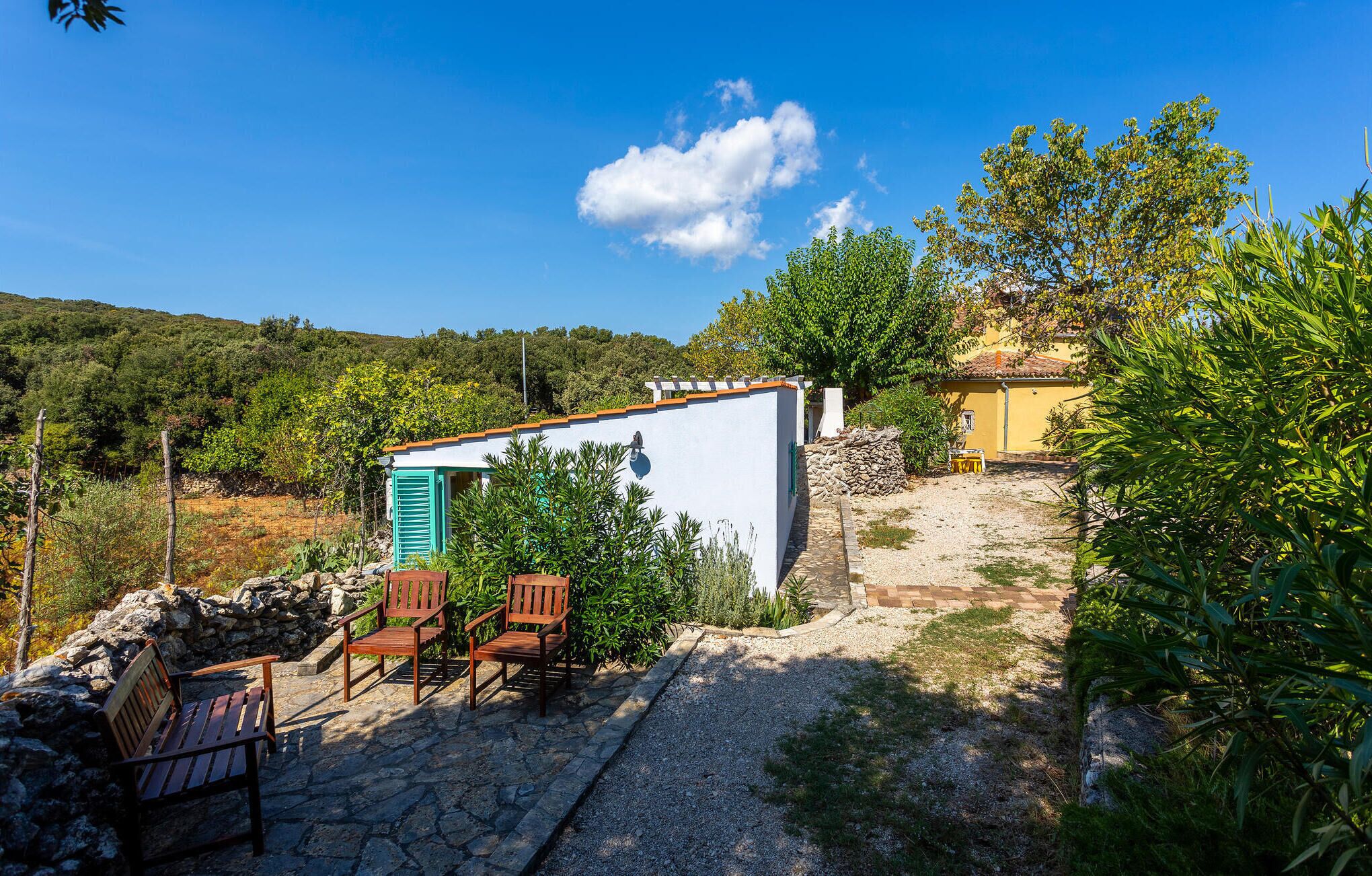 Un’area salotto esterna rustica con un tavolo in pietra e sedie di legno, un piccolo edificio bianco con persiana blu, e un edificio giallo sullo sfondo.