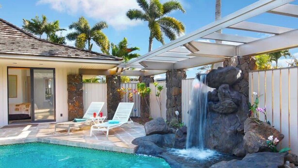 Una piscina con una cascada, sillas de descanso y un patio cubierto con palmeras al fondo.