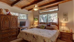 Un dormitorio con techo de madera, una cama con colcha estampada, un televisor sobre un mueble de madera y una ventana con vistas a árboles.