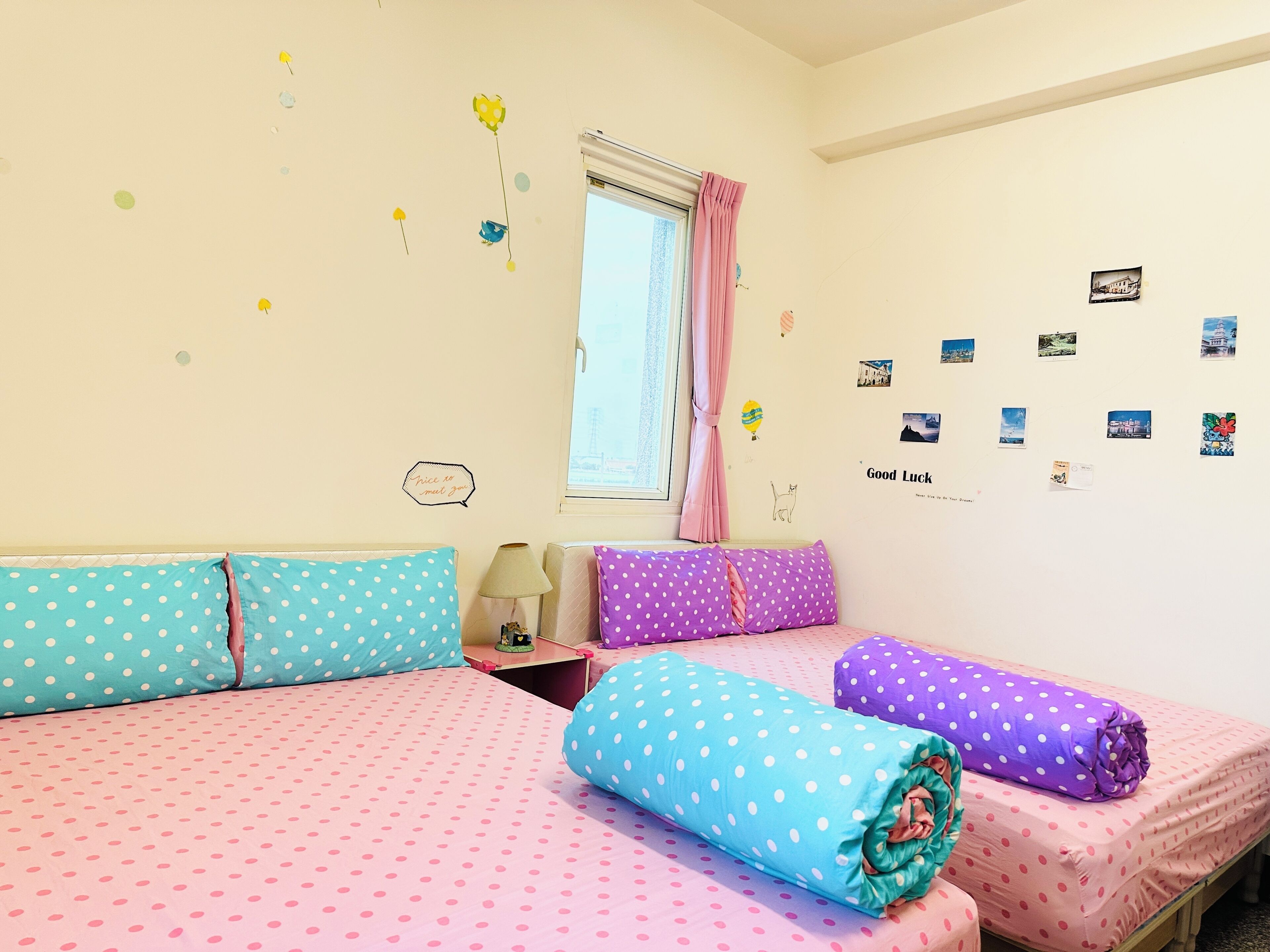 Una habitación con dos camas, cada una con ropa de cama de lunares en rosa y azul. Hay adornos en la pared, incluyendo un cartel que dice 'Good Luck' y fotografías pequeñas.