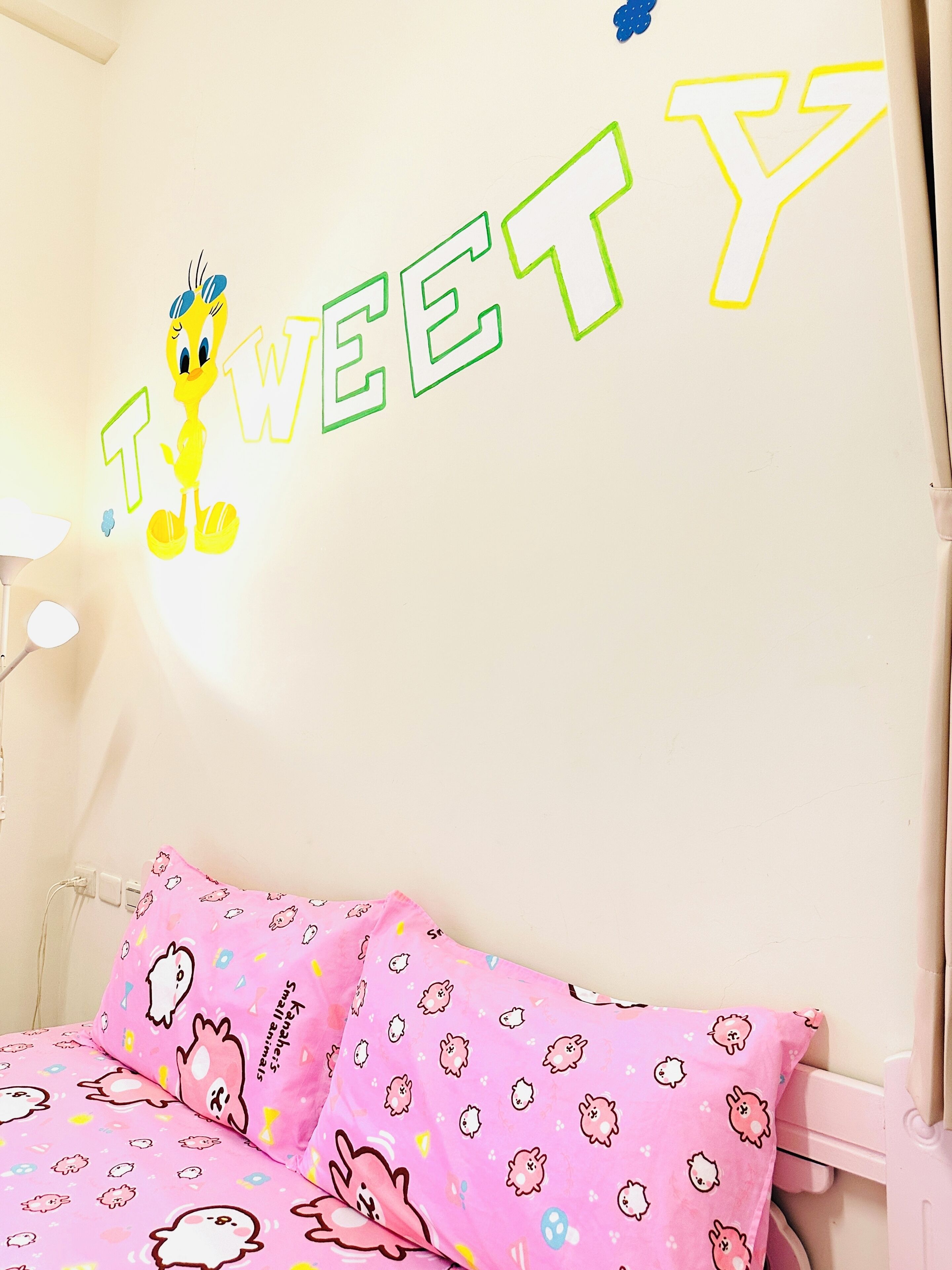 A bedroom with a bed featuring pink bedding with cartoon characters, a wall with the word "TWEETY" and a cartoon bird, and a white lamp.