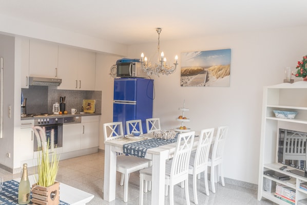 Une cuisine compacte avec des meubles blancs, un réfrigérateur bleu et un coin repas comprenant des chaises et une table, le tout dans des tons blancs.