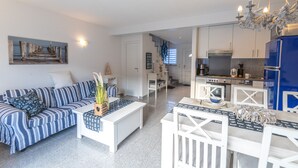A modern living room with a striped sofa, a white coffee table, and a dining area with white chairs. A kitchen with blue cabinets and a chandelier is visible in the background. - Vacation home on the Bodden near Fischland-Darß-Zingst; fenced-in garden. Dogs Welcome (Pruchten)