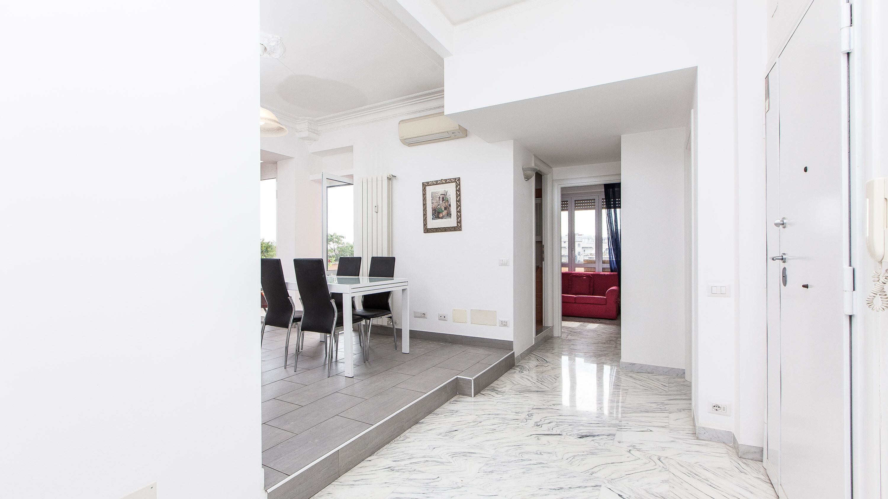 Un corridoio con pavimento in marmo che conduce a una sala da pranzo con tavolo e sedie bianche, un divano rosso sullo sfondo e una porta bianca a destra.