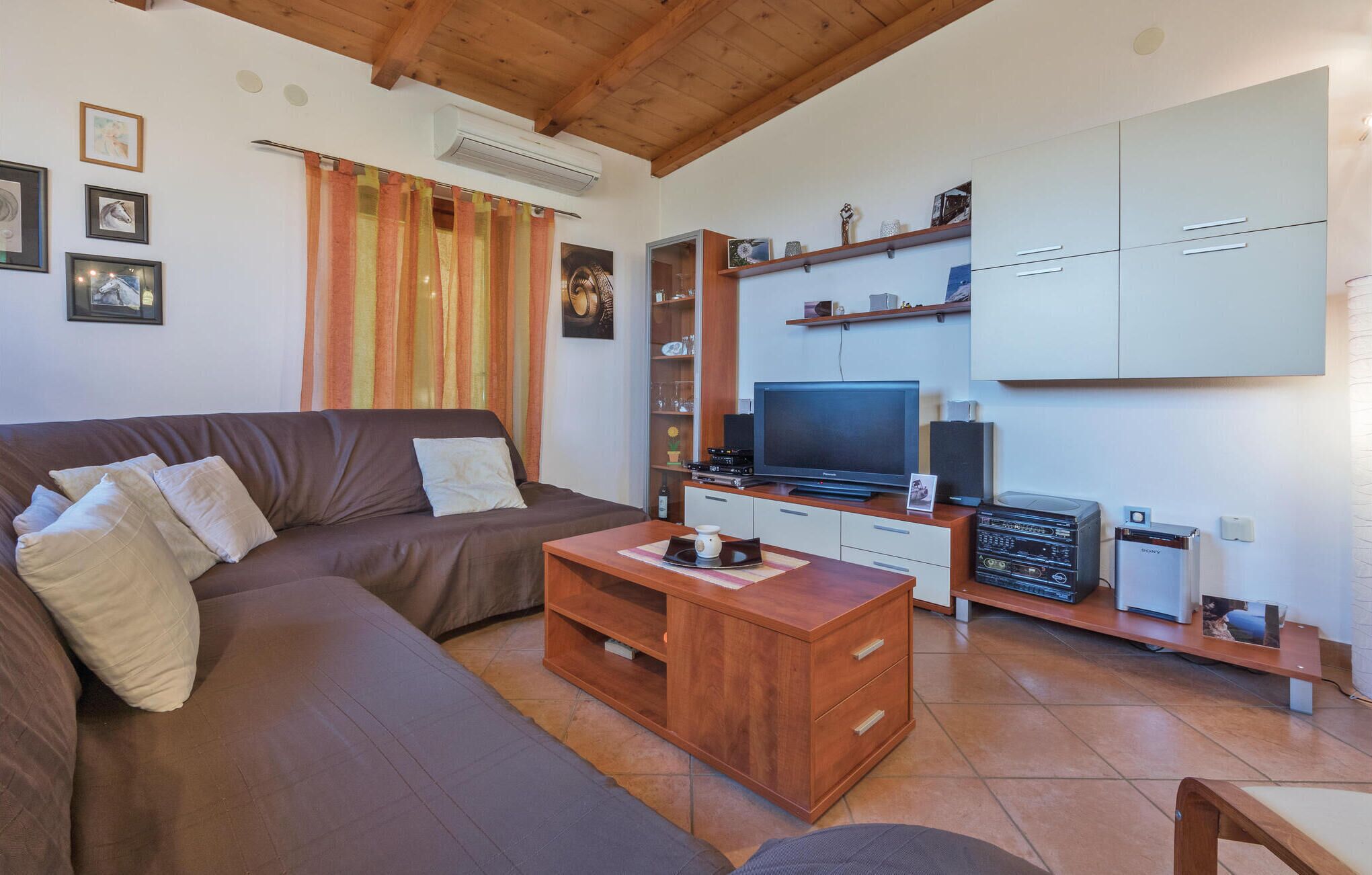 Una sala de estar con un sofá marrón, una mesa de centro de madera y un mueble de TV con televisor de pantalla plana, reproductor de DVD y altavoces.