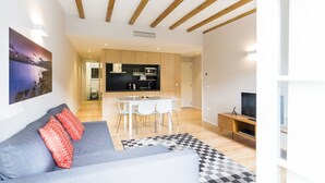Ein modernes Wohnzimmer mit einem grauen Sofa, roten Kissen, einem schwarz-weißen Schachbrettteppich, einer Holz-TV-Konsole und einer Küchenzeile mit Esstisch und Stühlen.
