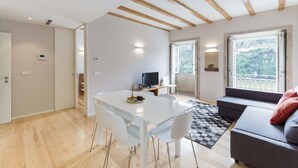 Ein modernes Wohnzimmer mit einem weißen Esstisch, einer grauen Couch und einem großen Fenster mit Blick auf Grünflächen.
