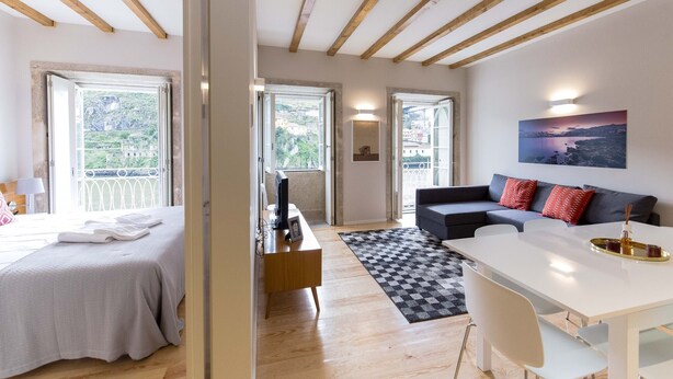 Ein modernes Zimmer mit Bett, Schreibtisch, Esstisch und einer Couch. Große Fenster bieten Blick ins Grüne.