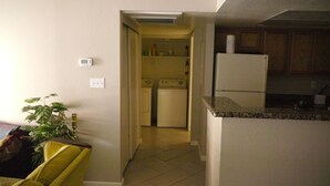 Une cuisine compacte équipée d’un réfrigérateur, d’un lave-linge et d’un four à micro-ondes. On aperçoit une plante et une chaise jaune au premier plan.