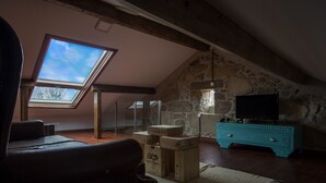 Een loft-achtige kamer met schuin dak, stenen muren, dakraam, een houten dressoir, een televisie en een leren bank.