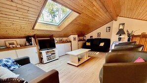 Un espace de vie chaleureux aménagé dans les combles, avec une lucarne, un plafond en bois, un téléviseur à écran plat, un canapé, une table basse et un coin repas.