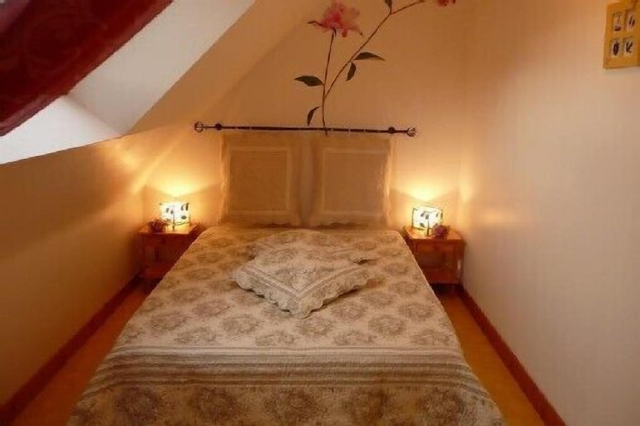 Une petite chambre à coucher accueillante, avec un lit simple, deux tables de chevet avec des lampes et une fleur décorative accrochée au mur.