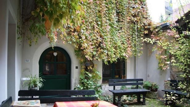 Ein Sitzbereich im Freien mit Tisch und Bänken, umgeben von viel Grün und einem weißen Gebäude mit grüner Tür.