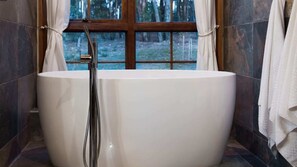 Una bañera independiente grande en un baño con vista a un bosque a través de una ventana.