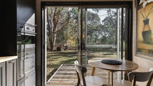 Una cuina moderna amb electrodomèstics d'acer inoxidable, zona de dinar amb taula rodona i cadires, i una porta corredissa de vidre que dona a una terrassa exterior amb vistes a arbres i un vehicle.