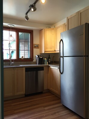 A kitchen with wooden cabinets, a stainless steel refrigerator, a dishwasher, and a window with a hanging light fixture.
