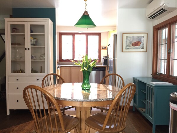 A dining area with a round table, four wooden chairs, a green vase with flowers, a cabinet with glass doors, and a kitchen in the background.