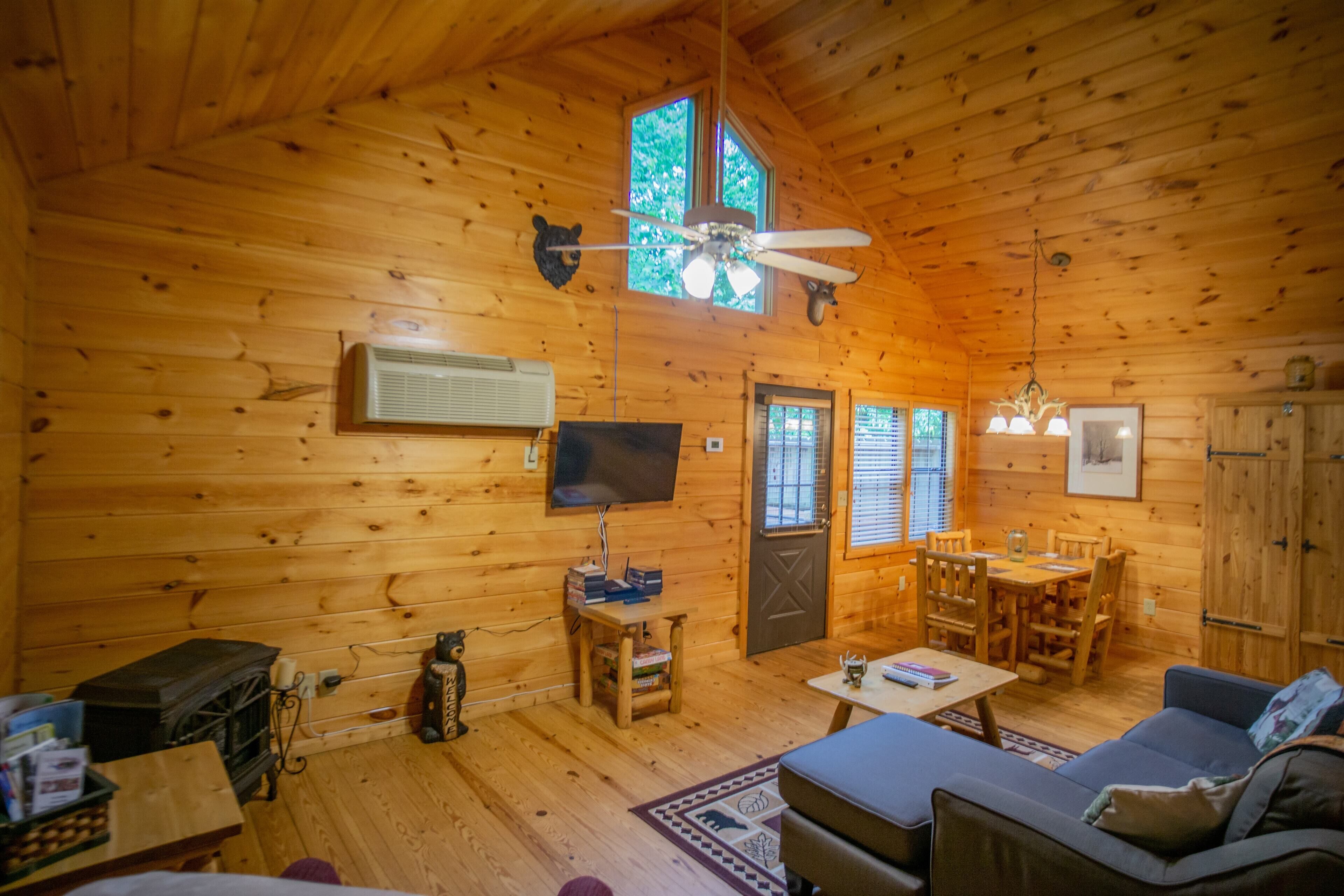 A cozy cabin interior with wooden walls, a ceiling fan, a television, a dining area, and a sofa.