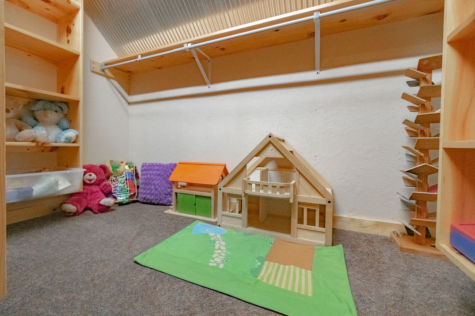 Une salle de jeux pour enfants avec une maisonnette en bois, des étagères remplies de jouets et un tapis de jeu vert.