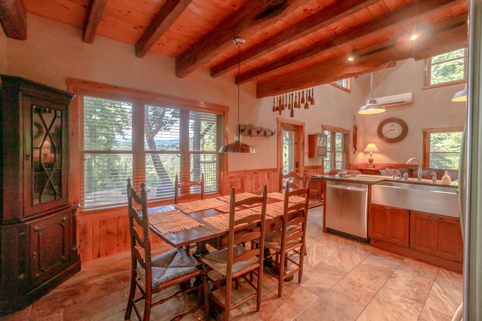 A rustic kitchen with wooden cabinets, a dining table with chairs, and a large window overlooking greenery.