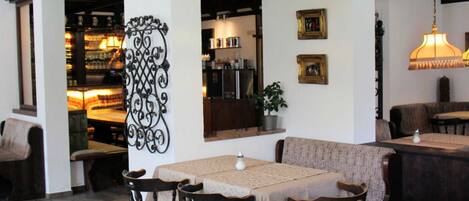 Un interior de restaurante con zona de bar, sillas de madera, mesas con manteles, lámparas colgantes y un elemento decorativo de metal en la pared.