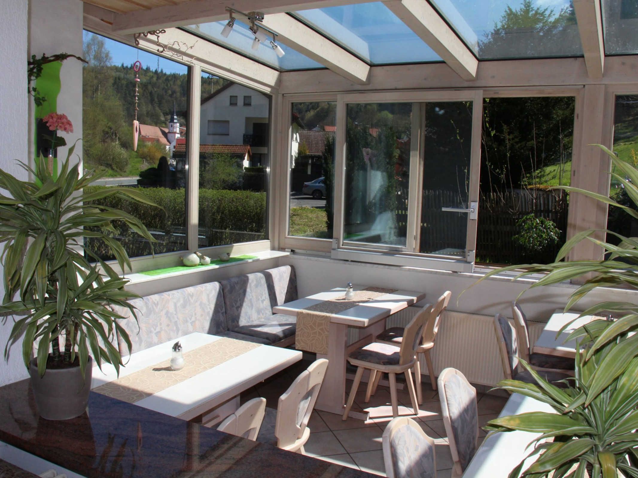A sunlit patio with a table and chairs, large windows, and potted plants.