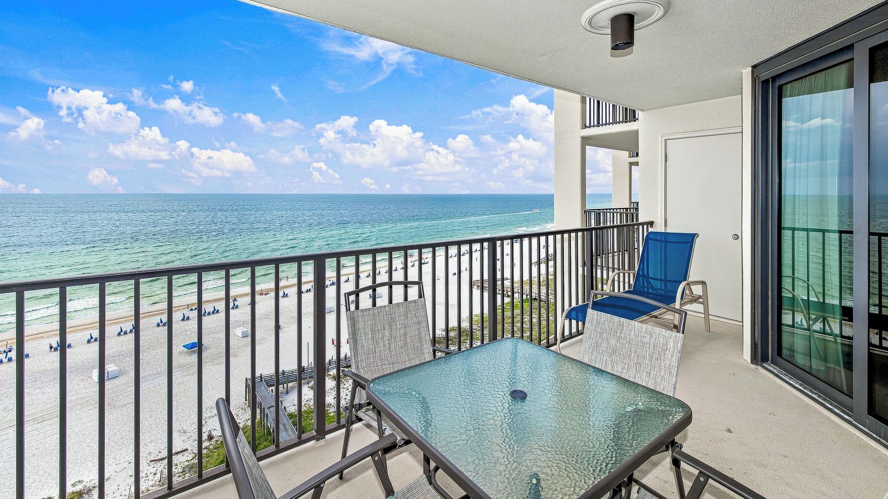 A balcony with a glass table and chairs, overlooking a beach with umbrellas and a clear sky.