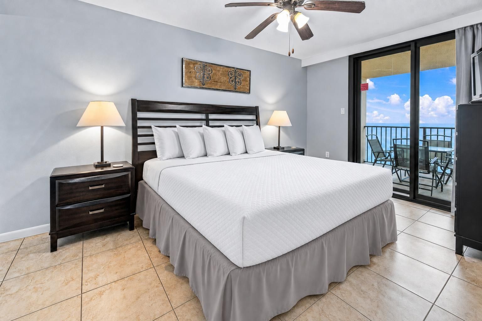 A bedroom with a large bed, two bedside tables with lamps, a ceiling fan, a balcony with a view, and a decorative wall piece.