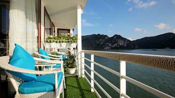 A balcony with wicker chairs and cushions, a potted plant, and a view of a lake with mountains in the background.