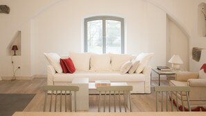 En stue med en hvid sofa, et lille bord og en stol. Et vindue viser udsigt til træer.