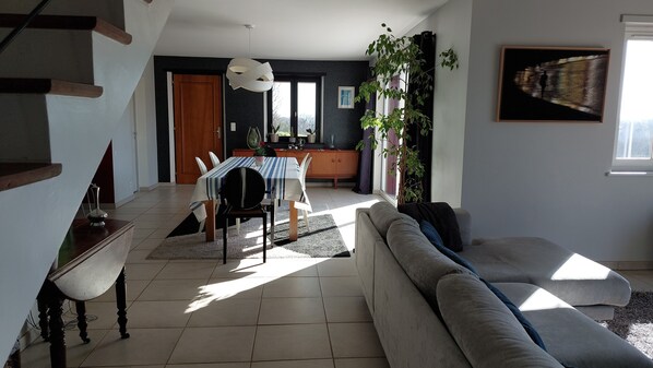 Ein modernes Wohnzimmer mit einer Couch, einem Esstisch und Stühlen. Eine Treppe befindet sich links, eine Pflanze und ein Fenster rechts.