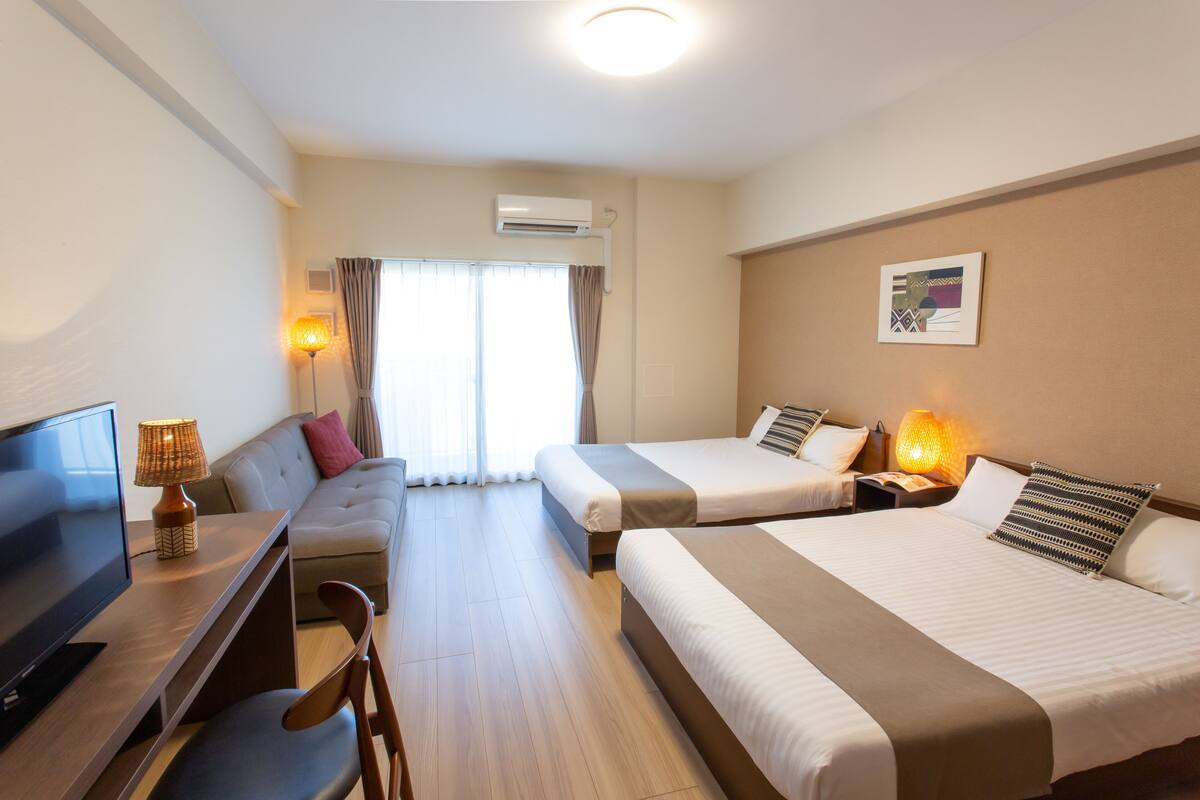 A hotel room with two beds, a desk with a TV, a sofa, and a window with curtains.