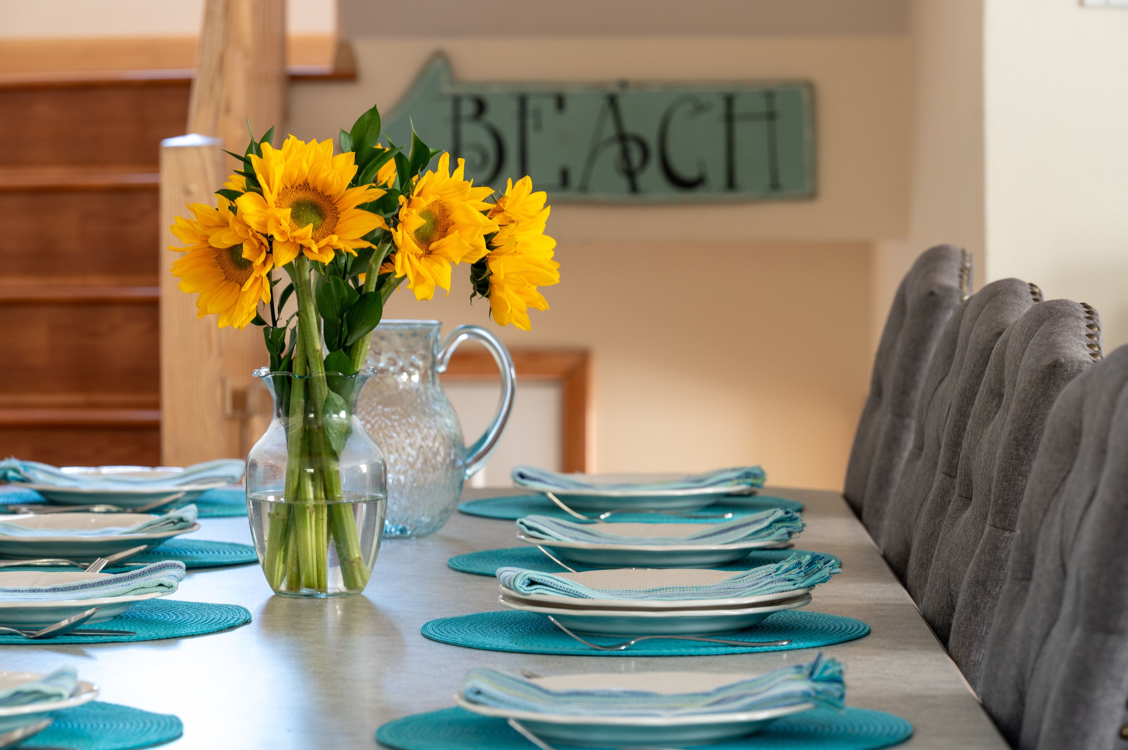 A dining table set with blue placemats and plates, a clear glass pitcher with water, and a vase with yellow flowers.