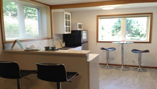 Een keuken met een magnetron, spoelbak en twee barkrukken. Er staan drie stoelen voor de keukenbar.