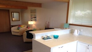 Een knus woongedeelte met een kitchenette, een bank, een lamp en een schilderij aan de muur.